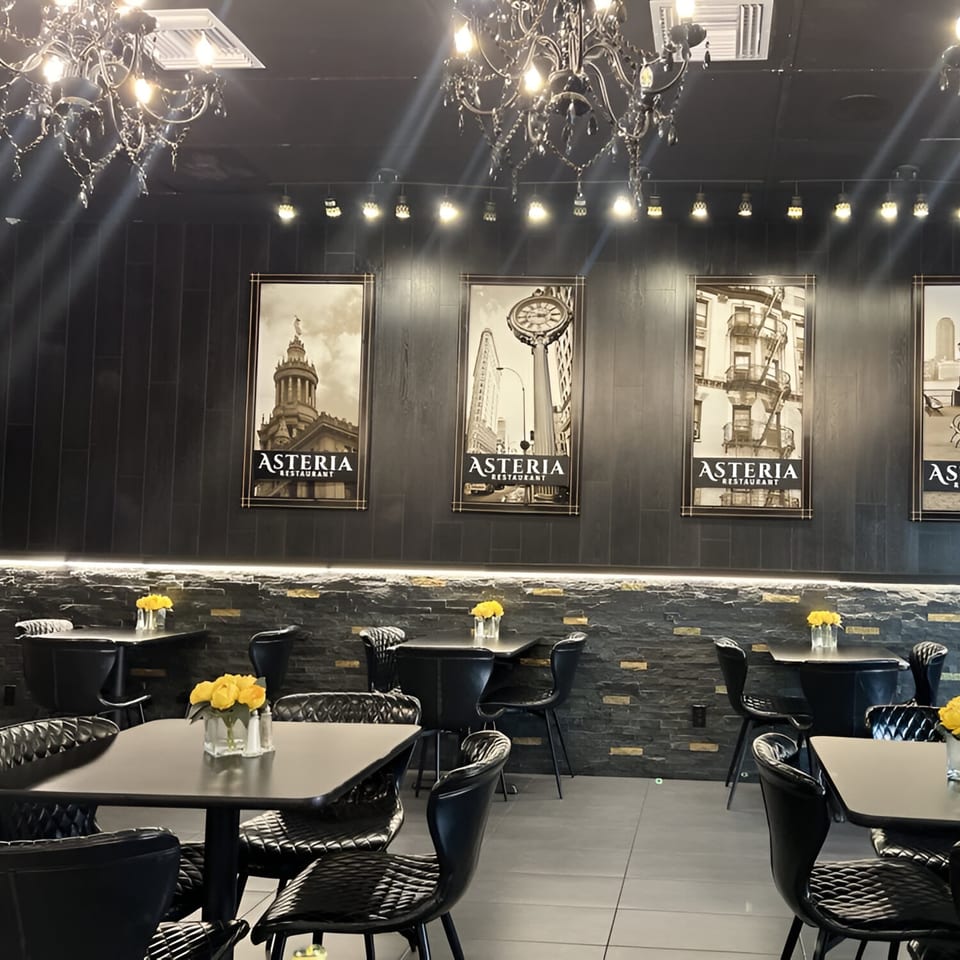 Interior of Asteria Restaurant with black tables, chairs, chandeliers, and framed city prints on dark walls.